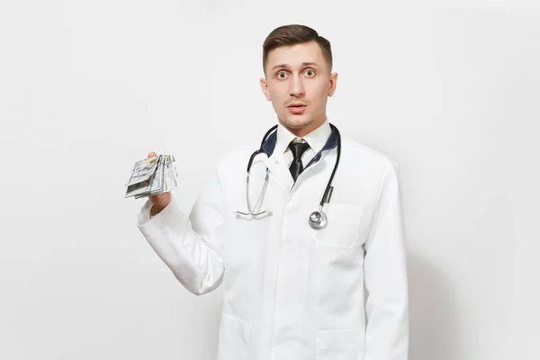 Shocked young doctor man isolated on white background. Male doctor in medical uniform, stethoscope holding bundle of dollars, banknotes, cash money. Healthcare personnel, health, medicine concept.