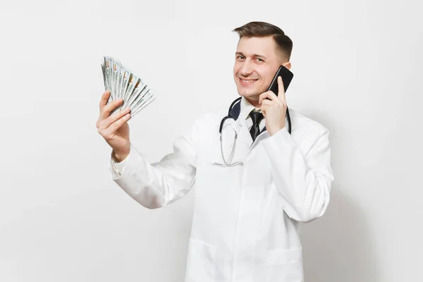 Усміхнений молодий лікар ізольований на білому тлі. Чоловік-лікар в медичній формі розмовляє на мобільному телефоні, тримає пакет доларів, банкноти, готівкові гроші. Медичний персонал, концепція медицини . — стокове фото