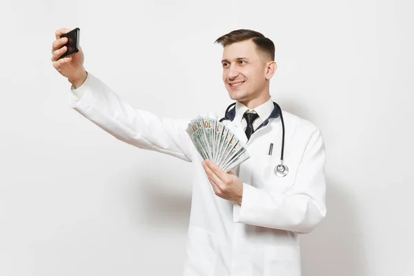 Усміхнений лікар чоловік ізольований на білому тлі. Чоловік-лікар у медичній формі робить селфі на мобільному телефоні, тримає пакет доларів, банкноти, готівкові гроші. Медичний персонал, концепція медицини . — стокове фото