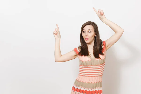 Hermosa joven morena en beige claro, vestido estampado rosa sonriendo, toma las manos en diferentes direcciones y señala sus dedos hacia arriba en el espacio de copia aislado sobre fondo blanco. Concepto de buen humor . — Foto de Stock