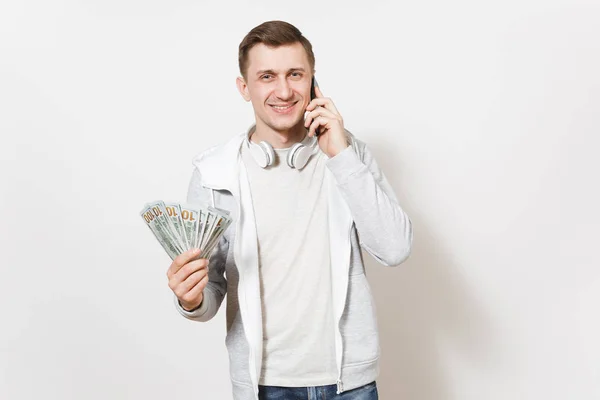 Junger gutaussehender lächelnder Mann im T-Shirt, helles Sweatshirt mit Kopfhörern um den Hals, der auf einem Handy mit einem Bündel Dollars spricht, Bargeld in der Hand isoliert auf weißem Hintergrund. Erfolgskonzept. — Stockfoto