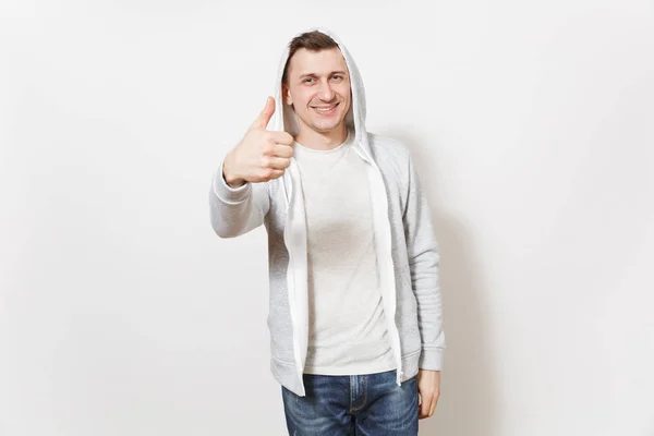 Young handsome smiling man in t-shirt, blue jeans and light sweatshirt shows gesture thumbs up and rejoices isolated on white background. Concept of emotions, good mood. Copy space for advertisement. — Stock Photo, Image