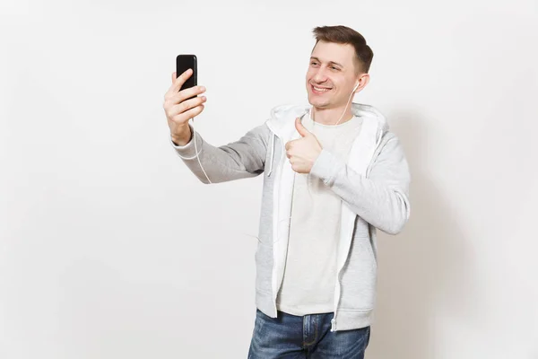年轻英俊的 t恤衫和轻型运动衫的男子在电话中自拍自己的手机, 并显示在工作室白色背景的手势大拇指。沟通理念, 心情好. — 图库照片
