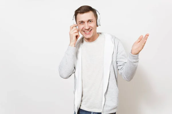 Joven estudiante hombre sonriente guapo en camiseta y sudadera ligera escuchando música con auriculares inalámbricos blancos y pone la mano a ellos aislados sobre fondo blanco. Concepto de emociones, estilo de vida —  Fotos de Stock