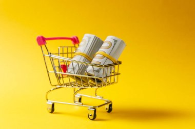 Supermarket grocery push cart for shopping with pink elements on handle with bundles of dollars money banknote cash isolated on yellow background. Concept of shopping. Copy space for advertisement. clipart
