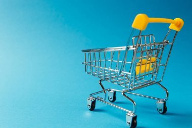 Süpermarket bakkal itme sepetinin siyah jantlar ve tutamacı üzerinde mavi arka plan izole sarı plastik öğelerle birlikte alışveriş için kapatın. Alışveriş kavramı. Kopya alanı reklam için