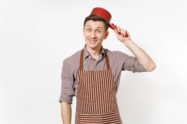 Young fun crazy happy man chef or waiter in striped brown apron, shirt holding red empty stewpan on head isolated on white background. Male housekeeper or houseworker. Kitchenware and cuisine concept.