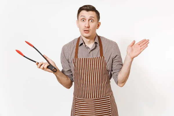 Joven chef con delantal marrón rayado, camisa con cocina negra que sirve un par de pinzas de plástico para ensalada aislada sobre fondo blanco. Hombre ama de llaves o empleada doméstica. Menaje de cocina, concepto de cocina . —  Fotos de Stock