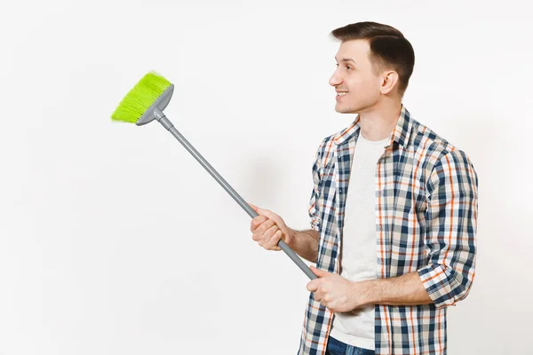 Joven ama de llaves con camisa a cuadros sosteniendo y barriendo con escoba verde aislada sobre fondo blanco. Hombre haciendo tareas domésticas, mirando a un lado. Copia espacio para la publicidad. Concepto de limpieza . — Foto de Stock