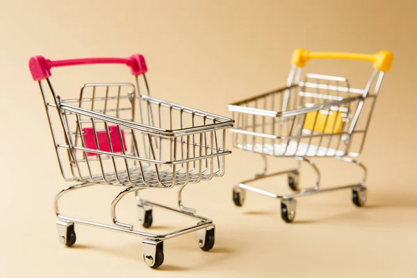 Close up of two supermarket grocery push carts for shopping with wheels and yellow and pink plastic elements on handle isolated on beige background. Concept of shopping. Copy space for advertisement