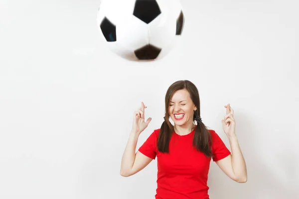 European young woman two fun pony tails, football fan or player in red uniform keep fingers crossed, flying soccer ball isolated on white background. Sport play football lifestyle concept. Make wish.