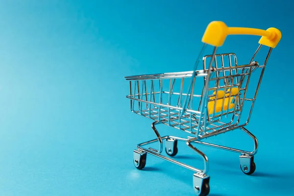 スーパー マーケット食料品は黒色車輪と青の背景に分離されたハンドルに黄色のプラスチック要素ショッピング カートを押すをクローズ アップ。ショッピングのコンセプト。広告のためのスペースをコピーします。 ストック画像