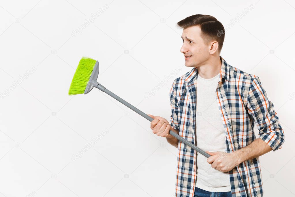 Young strange fun housekeeper man in checkered shirt holding and sweeping with green broom isolated on white background. Male doing house chores. Copy space for advertisement. Cleanliness concept.