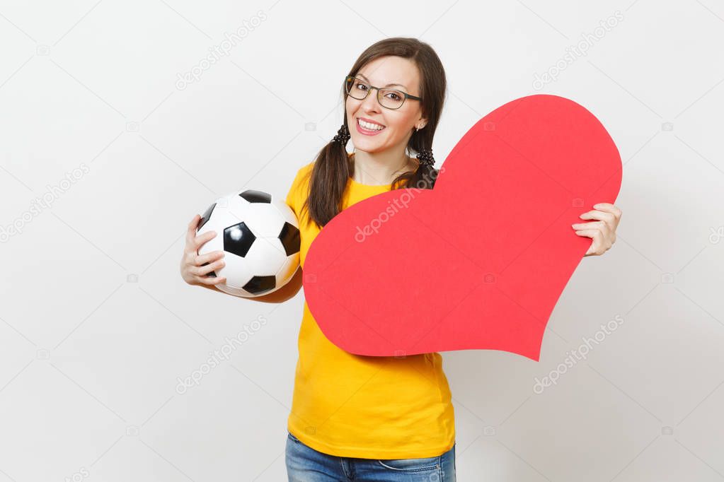 Smiling European woman, fun pony tails, football fan or player in glasses, yellow uniform hold classic soccer ball, red heart isolated on white background. Sport, football, healthy lifestyle concept.
