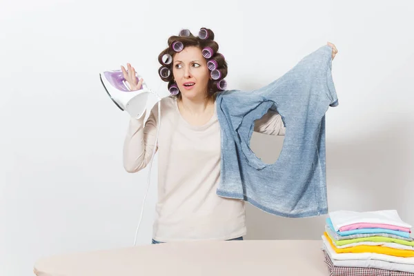 Chocado diversão louca dona de casa, frisadores no cabelo em roupas leves segurando camisa queimada com buraco feito de ferro, de pé na tábua de engomar. Mulher isolada sobre fundo branco. Espaço de cópia para propaganda . — Fotografia de Stock