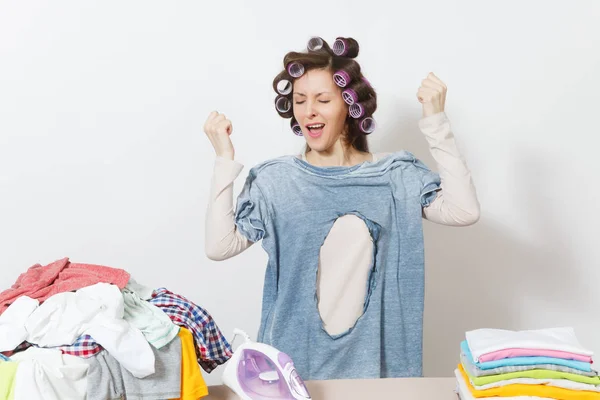 Scioccato divertimento folle casalinga, bigodini sui capelli in abiti leggeri in possesso di camicia bruciata con foro fatto da ferro, in piedi a asse da stiro. Donna isolata su sfondo bianco. Copia spazio per pubblicità . — Foto Stock