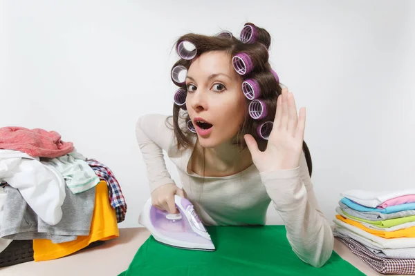 Close up divertente casalinga con bigodini sui capelli in abiti leggeri stand a asse da stiro con ferro da stiro, abbigliamento di famiglia. Donna origliare, udito gesto isolato su sfondo bianco. Copia spazio annuncio . — Foto Stock