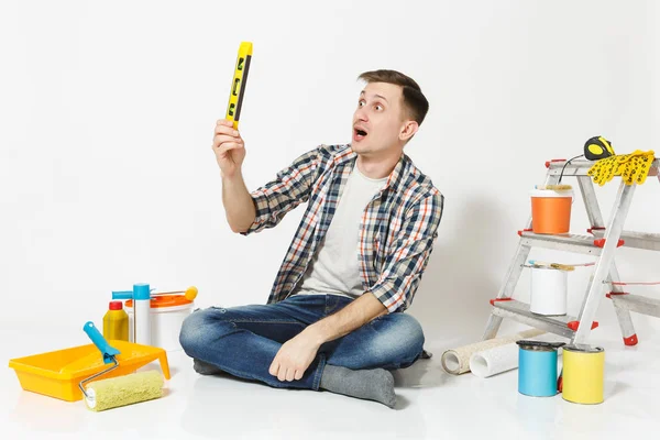 Man in casual clothes sitting on floor with building bubble spirit level, instruments for renovation apartment isolated on white background. Wallpaper, gluing accessories, tools. Repair home concept.