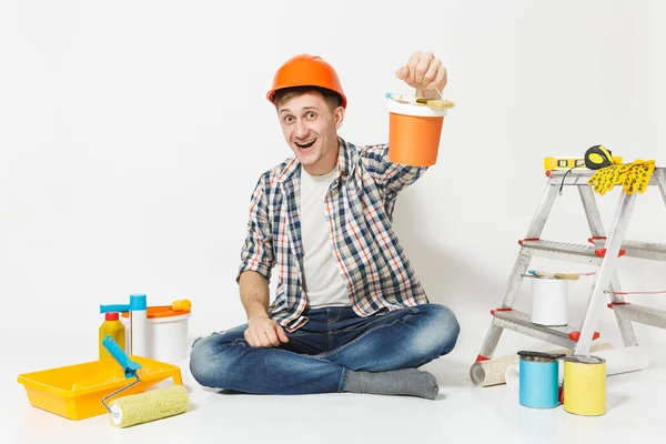 Homem de capacete de proteção laranja sentado no chão com lata de tinta, instrumentos para renovação apartamento isolado no fundo branco. Papel de parede, acessórios de colagem, ferramentas de pintura. Reparação home concept . — Fotografia de Stock
