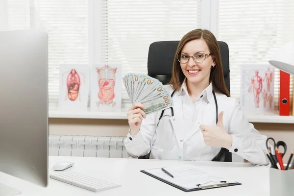 Young smiling female doctor sitting at desk holding bundle of dollars cash money, working with medical documents in light office in hospital. Woman in medical gown in consulting room. Medicine concept
