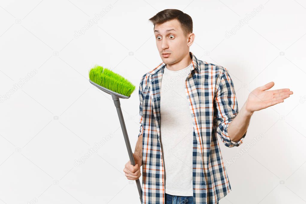 Young strange fun housekeeper man in checkered shirt holding and sweeping with green broom isolated on white background. Male doing house chores. Copy space for advertisement. Cleanliness concept.