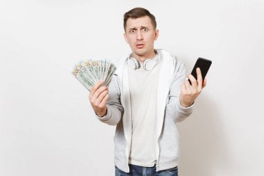 Yakışıklı delikanlı t-shirt ve boynuna kulaklıklarla ışık kazak dolar, nakit para, cep telefonu ve beyaz arka plan üzerinde izole şaşkınlık elinde tutar. Başarı kavramı