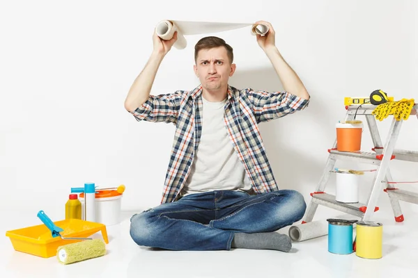 Homem perplexo confuso sentado no chão com rolo de papel de parede acima da cabeça, instrumentos para renovação apartamento isolado no fundo branco. Acessórios de colagem, ferramentas de pintura. Reparação home concept . — Fotografia de Stock