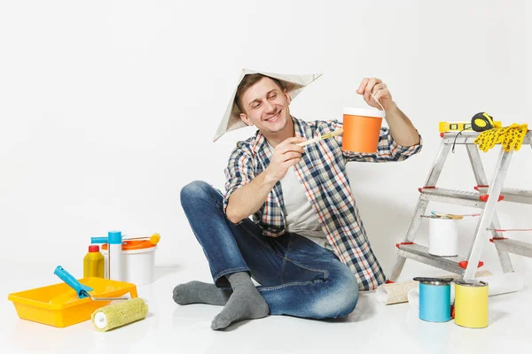 Homem de chapéu de jornal sentado no chão com pincel, lata, instrumentos para renovação apartamento quarto isolado no fundo branco. Papel de parede, acessórios de colagem, ferramentas de pintura. Reparação home concept . — Fotografia de Stock