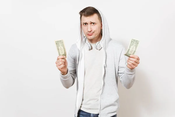 Giovane studente bel ragazzo in t-shirt, jeans blu e felpa leggera con cappuccio con cuffie tiene due banconote da un dollaro e sembra perplesso isolato su sfondo bianco. Concetto di emozioni — Foto Stock