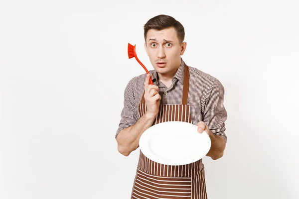 Młody człowiek szefa kuchni w paski brązowy fartuszek, koszula posiada myje biały okrągły pusty płyta jasne czerwone szczotką do mycia naczyń na białym tle. Mężczyzna gospodyni, houseworker lub pracownik domowy — Zdjęcie stockowe