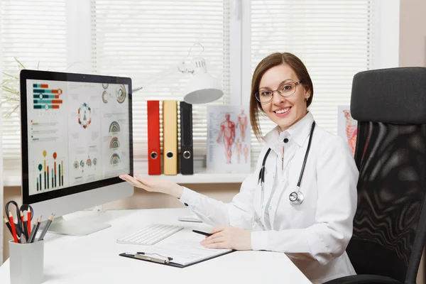 Mladá usměvavá žena sedí u stolu, pracovat na počítači s lékařské dokumenty ve světle kanceláři v nemocnici. Lékařka v lékařské šaty, stetoskop v ordinaci. Zdravotnictví, lékařství koncepce — Stock fotografie