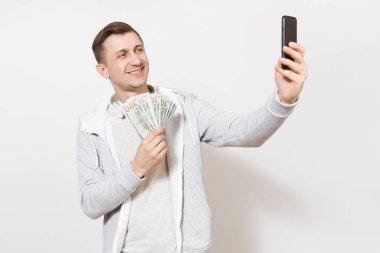 Genç gülümseyen yakışıklı t-shirt, boynuna kulaklıklarla ışık kazak bekle paket dolar, nakit para ve resimlerin kendini beyaz arka plan üzerinde izole telefon. Başarı kavramı