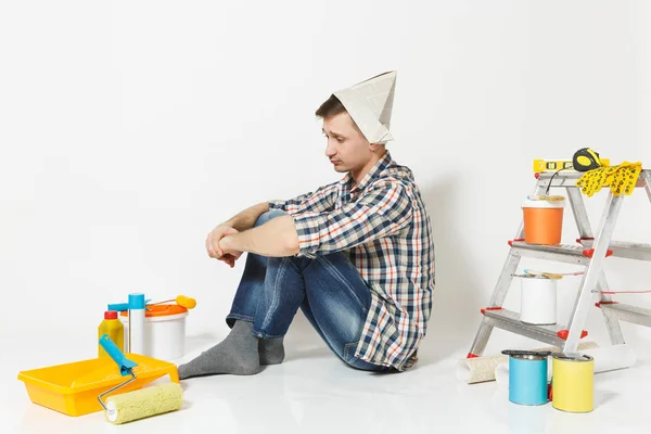 Νεαρός άνδρας με το καπέλο εφημερίδα κάθεται στο πάτωμα με τα όργανα για δωμάτιο διαμέρισμα ανακαίνιση που απομονώνονται σε λευκό φόντο. Ταπετσαρία, κόλλημα αξεσουάρ, εργαλεία βαφής. Επισκευή concept home. Πλάγια όψη. — Φωτογραφία Αρχείου