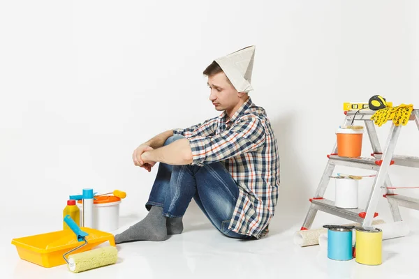 Joven en sombrero de periódico sentado en el suelo con instrumentos para la renovación habitación de apartamentos aislado sobre fondo blanco. Fondo de pantalla, pegar accesorios, herramientas de pintura. Reparar concepto de casa. Vista lateral . — Foto de Stock