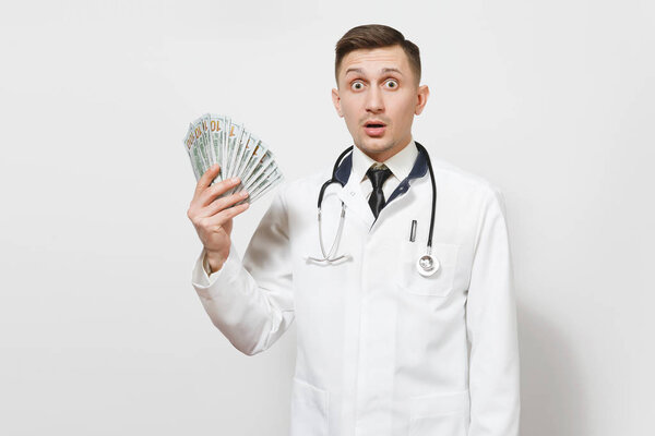 Shocked young doctor man isolated on white background. Male doctor in medical uniform, stethoscope holding bundle of dollars, banknotes, cash money. Healthcare personnel, health, medicine concept.