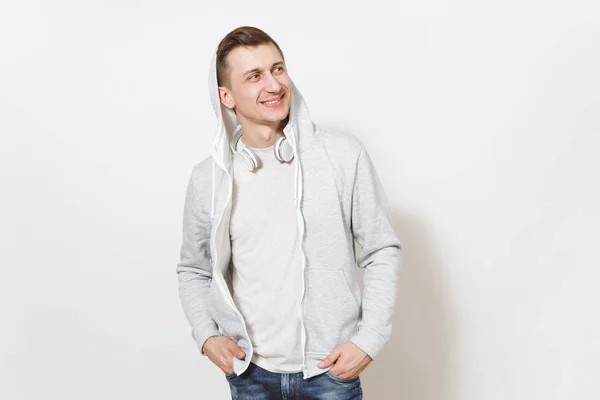 Νέων όμορφος άντρας χαμογελώντας σε μπλε τζιν, t-shirt και ελαφρύ φούτερ με κουκούλα με ακουστικά κρατά τα χέρια στις τσέπες του και αναζητούν μακριά στο studio σε λευκό φόντο. Αντίληψη των συναισθημάτων — Φωτογραφία Αρχείου
