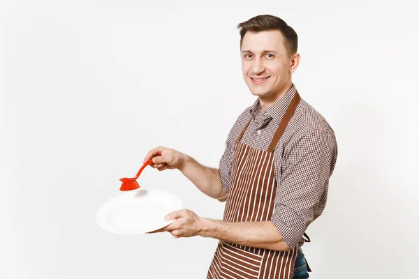 Jonge man chef-kok in gestreepte bruine schort, shirt houdt wast witte ronde lege duidelijk plaat met rode borstel voor afwas geïsoleerd op een witte achtergrond. Mannelijke huishoudster, houseworker of huishoudelijk werker — Stockfoto