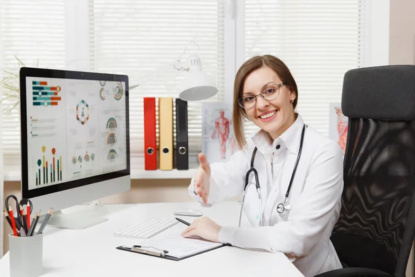 Mladá usměvavá žena sedí u stolu, pracovat na počítači s lékařské dokumenty ve světle kanceláři v nemocnici. Lékařka v lékařské šaty, stetoskop v ordinaci. Zdravotnictví, lékařství koncepce — Stock fotografie