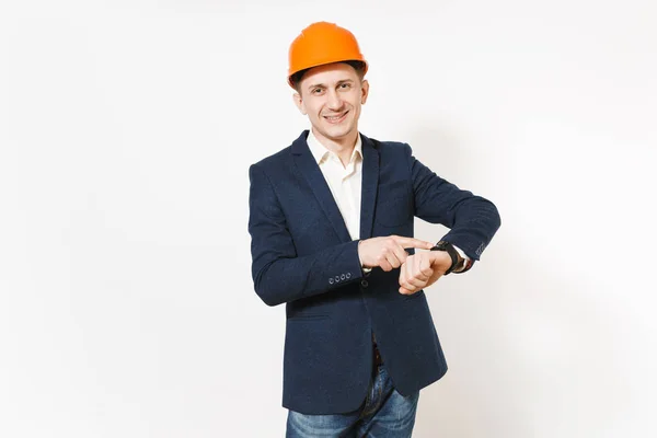 Jovem homem de negócios sorridente em terno escuro, construção protetora capacete laranja apontando dedo indicador no relógio na mão isolado no fundo branco. O tempo está a acabar. Trabalhador masculino para publicidade . — Fotografia de Stock