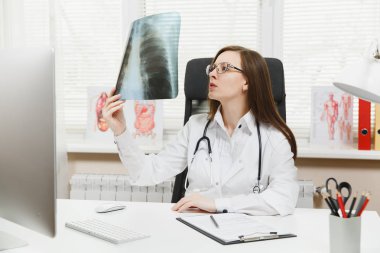 Female doctor sitting at desk, working on computer, holding X-ray of lungs, fluorography, roentgen in light office in hospital. Woman in medical gown, stethoscope in consulting room. Medicine concept. clipart