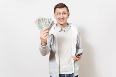 Genç yakışıklı gülümseyen adam öğrenci tişört, beyaz arka plan üzerinde izole elinde bohça-in dolar, nakit para, cep telefonu boynuna kulaklıklarla ışık kazak tutar. Başarı kavramı.