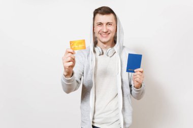 Genç yakışıklı gülümseyen mutlu adam t-shirt, kulaklık ile hood ile hafif kazak beyaz arka plan üzerinde izole elinde uluslararası pasaport ve kredi kartı tutar. Seyahat, turizm kavramı