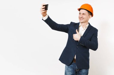 Genç yakışıklı gülümseyen iş adamı koyu takım, koruyucu hardhat başparmak gösterilmesini ve beyaz arka plan üzerinde izole cep telefonunda selfie yapıyor. Erkek işçi reklam için. İş kavramı.