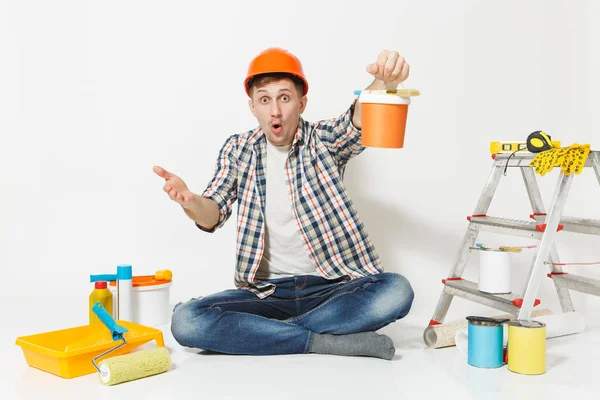 Homem chocado em capacete de proteção laranja sentado no chão com lata de tinta, instrumentos para renovação apartamento isolado no fundo branco. Papel de parede, acessórios, ferramentas de pintura. Reparação home concept . — Fotografia de Stock