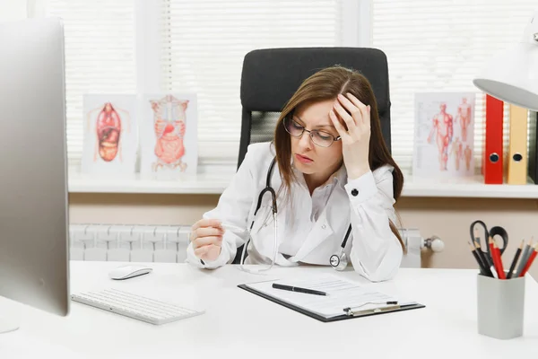 Female doctor sitting at desk with medical documents, clinical thermometer, high fever temperature in light office in hospital. Woman in medical gown, stethoscope in consulting room. Medicine concept.