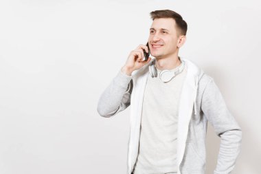 Genç yakışıklı gülümseyen adam öğrenci tişört ve boynuna kulaklıklarla ışık kazak cep telefonu üzerinde görüşmeler ve izole üzerinde beyaz arka plan sevinir. İletişim, duyguların kavramı.
