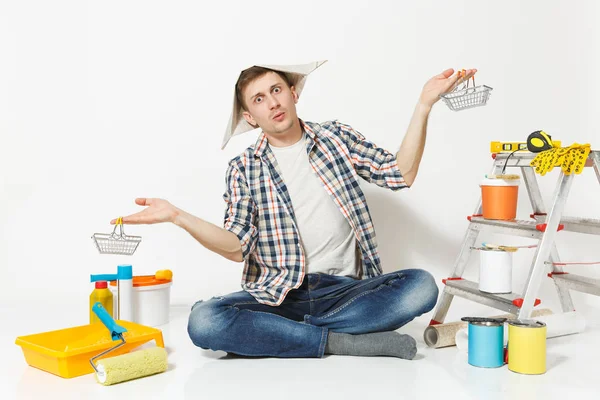 Hombre divertido en sombrero de periódico sostiene cestas de supermercado de metal para ir de compras. Instrumentos para la renovación apartamento aislado sobre fondo blanco. Fondos de pantalla, accesorios, herramientas de pintura. Concepto de reparación — Foto de Stock