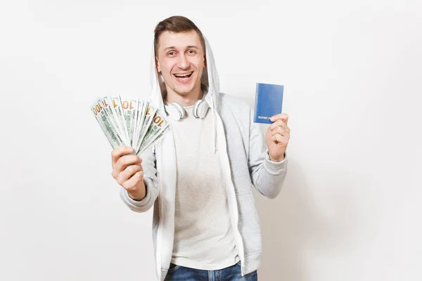 Jonge knappe Glimlachende man in t-shirt, lichte sweatshirt met capuchon met koptelefoon houdt internationaal paspoort, bundel van dollar contant geldgeld geïsoleerd op een witte achtergrond. Concept van reizen, toerisme. — Stockfoto