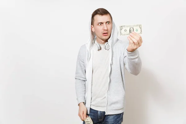 Jonge knappe geschokt man in t-shirt en lichte sweatshirt met capuchon met koptelefoon houdt één-dollarbiljetten en verbaasde blikken bij hen geïsoleerd op een witte achtergrond. Concept van emoties — Stockfoto