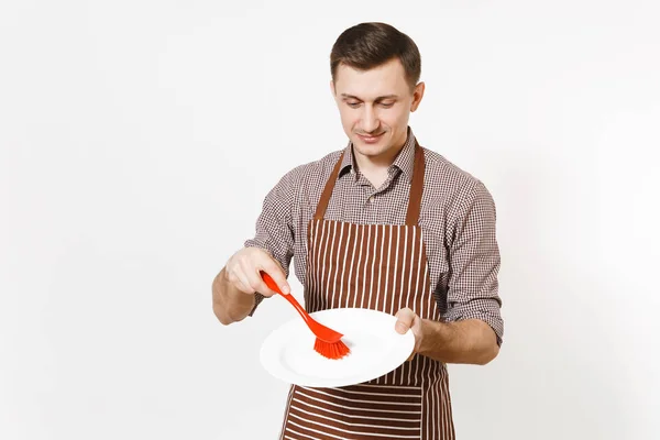 Młody człowiek szefa kuchni w paski brązowy fartuszek, koszula posiada myje biały okrągły pusty płyta jasne czerwone szczotką do mycia naczyń na białym tle. Mężczyzna gospodyni, houseworker lub pracownik domowy — Zdjęcie stockowe